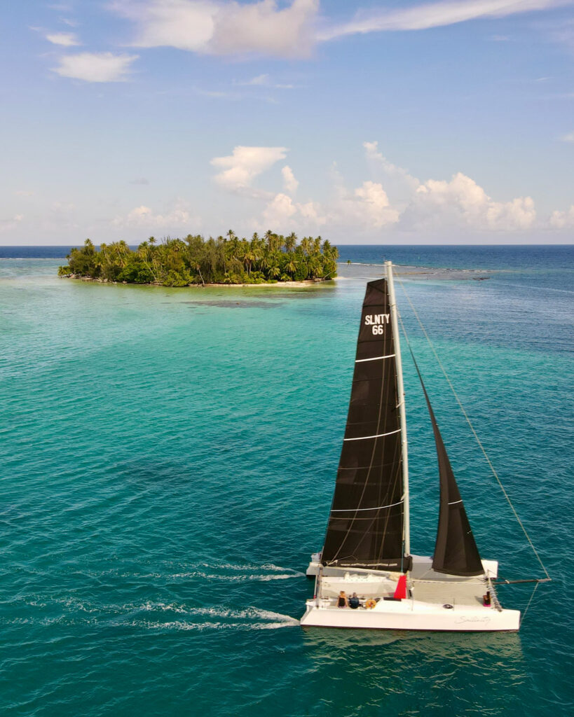 Catamaran sailing toward a palm-covered tropical island with turquoise water and blue sky above tall white clouds.