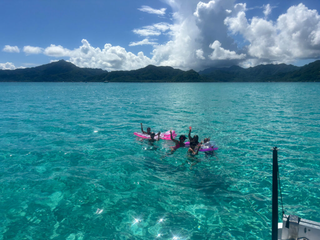 A group of people in pink flotation devices enjoying clear turquoise seawater near a mountainous coastline under a partly cloudy sky