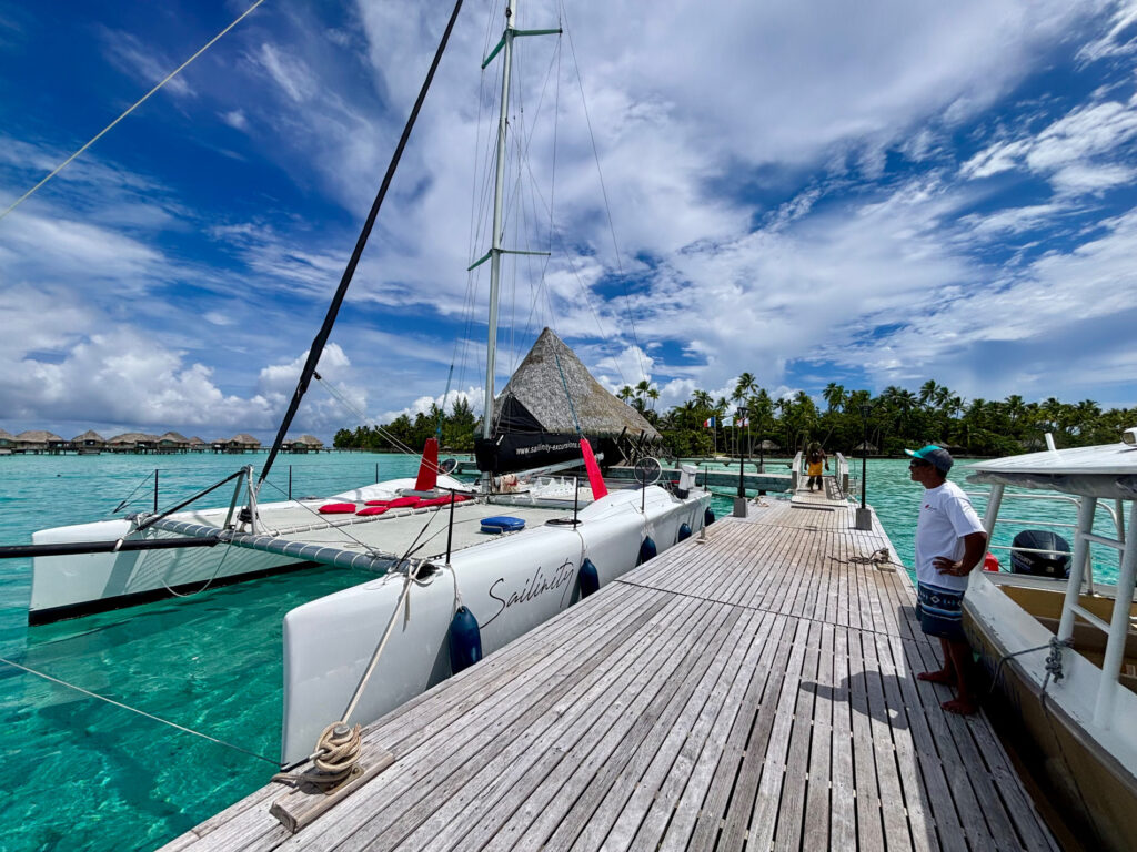 Catamaran moored at a wooden dock in turquoise tropical water, with palm trees and a thatched hut in the background.