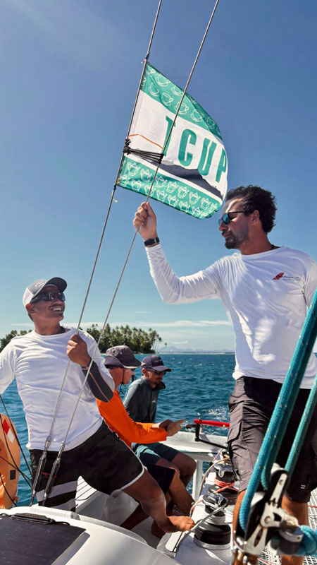 l-histoire-derriere-sailinity Two men on a sailboat raise a teal-and-white flag as the bright sky and ocean surround them, with other crew in the background.