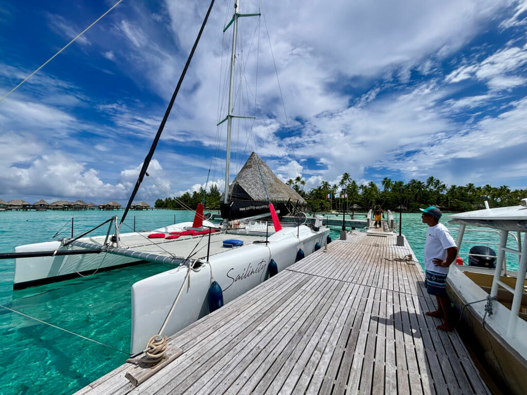 le-66-devenu-sailinity Sailboats docked at a wooden pier beside turquoise water under a blue sky with palm trees in the background, a man standing at right.