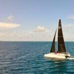 Catamaran with black sails labeled SLNTY 66 sailing on calm blue ocean under a clear sky.