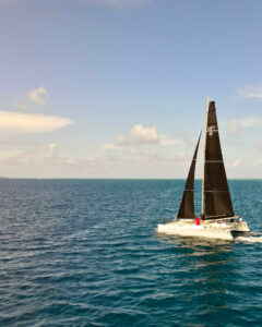 Catamaran with black sails labeled SLNTY 66 sailing on calm blue ocean under a clear sky.
