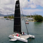 Catamaran with a tall black mainsail labeled SLNTY 66 in a marina, with people on deck and a small dinghy nearby.