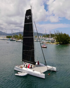 Catamaran with a tall black mainsail labeled SLNTY 66 in a marina, with people on deck and a small dinghy nearby.