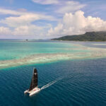 A sailing catamaran with black sails glides over turquoise water toward a forested island with rolling hills under a partly cloudy sky.