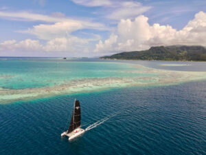 A sailing catamaran with black sails glides over turquoise water toward a forested island with rolling hills under a partly cloudy sky.