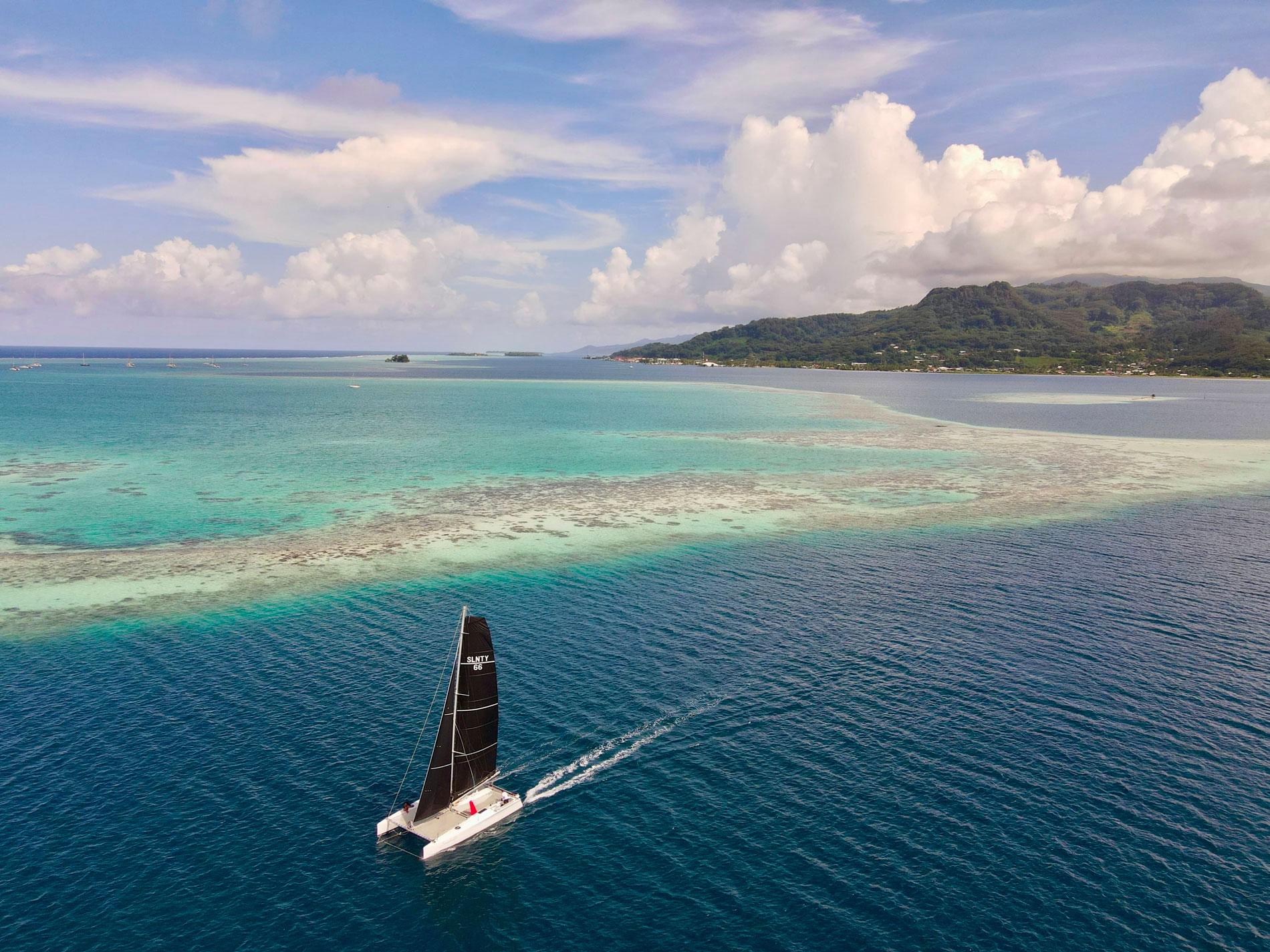 A sailing catamaran with black sails glides over turquoise water toward a forested island with rolling hills under a partly cloudy sky.